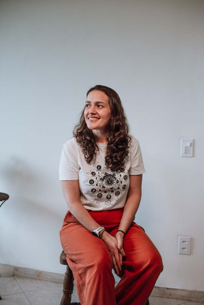 Paula Abrantes sentada sorrindo, com camiseta branca e calça laranja